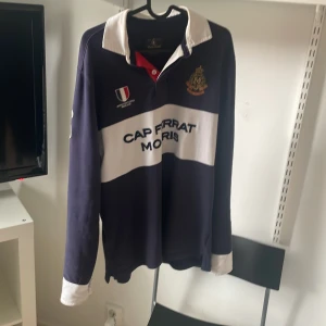 Morris pikétröja med brodyr - Säljer en långärmad pikétröja från Morris i mörkblått och vitt med broderad logga och texten 'Cap Ferrat'. Klassisk vit krage och manschetter, samt snygga detaljer i rött vid knäppningen. Perfekt för en preppy look.