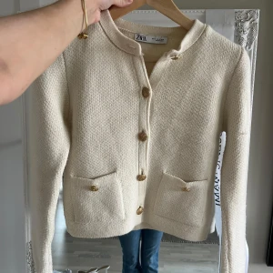 Beige kofta från Zara med guldknappar - Superfin beige kofta från Zara med snygga guldknappar framtill och två fickor. Koftan är stickad, har rund halsringning och långa ärmar. Perfekt för lager-på-lager och ger en clean och stilren look.