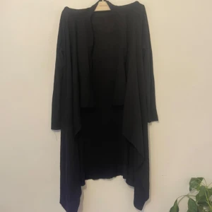 Svart lång kofta med öppen front - Svart lång kofta med öppen front och draperad nederkant. Koftan har lång ärm och är tillverkad i ett tunt, stickat material som ger en snygg och avslappnad look. Perfekt att slänga över en t-shirt eller topp för extra lager. Knappt använt.