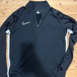 Nike svart Dri-Fit half zip tröja - Svart långärmad Nike Dri-Fit tröja med vit logga på bröstet och vita paneler längs sidorna. Tröjan har en hög krage och half zip-dragkedja framtill. Perfekt för träning eller sportiga outfits. Passar S och M med lite tightare passform. Kan kombineras med shortsen för bättre pris.                                                                              MVH
