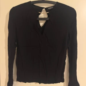 Svart långärmad blus från Mango - Svart långärmad blus från Mango Basics med v-ringning och dekorativt metallspänne vid halsen. Tunn och lätt i materialet, perfekt för en stilren look. Blusen har enkla muddar med knappdetalj och en rak passform.