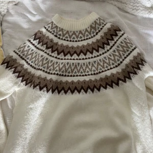 Vit stickad tröja med brunt mönster - Supermysig vit stickad tröja med klassiskt nordiskt mönster i bruna och beige toner över axlar och bröst. Tröjan har rund halsringning och långa ärmar, perfekt för kalla dagar. Snygg att styla med jeans eller kjol för en chill vinterlook.