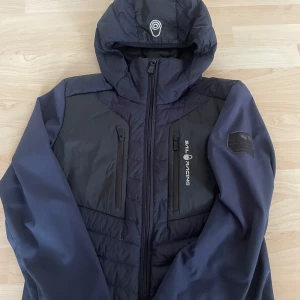 Sail racing jacka som ny - Denna Sail Racing-jacka är en stilren och funktionell ytterjacka, perfekt för både vardag och äventyr. Jackan har en marinblå och svart färgkombination som ger ett sportigt och modernt utseende. Den är utrustad med en praktisk huva som skyddar mot vind och regn, samt en smidig dragkedja framtill för enkel av- och påklädning. Två bröstfickor ger gott om förvaring för småsaker. På bröstet finns den välkända Sail Racing-loggan, och på vänster ärm syns Antarctic-loggan, vilket understryker jackans 