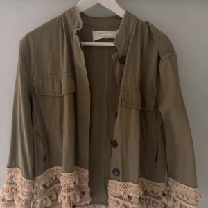Olivgrön jacka med virkade detaljer Zara - Olivgrön jacka från Zara med snygga virkade och fransade detaljer i beige längs ärmslut och nederkant. Jackan har stora bröstfickor, knappar framtill och en rak, loose passform. Perfekt för dig som vill sticka ut med coola detaljer.