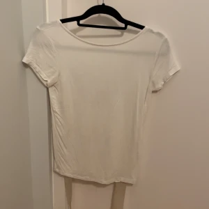 Vit topp med öppen rygg och knytband - Säljer en vit kortärmad topp med rund halsringning fram och djup öppen rygg med knytband. Toppens enkla design gör den lätt att styla och den är tillverkad i mjukt bomullsmaterial. Perfekt för dig som gillar minimalistisk stil med en twist.