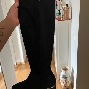 Svarta höga stövlar med gulddetalj - Snygga svarta höga stövlar i mockaimitation med rund tå och låg klack. Dragkedja på sidan guldfärgad detalj på hälen. Storlek 39, aldrig användts utomhus.                                                                              (Läs alltid min profil för villkor)