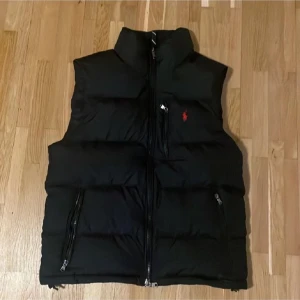 Svart dunväst från Polo Ralph Lauren - Svart dunväst från Polo Ralph Lauren med hög krage och dragkedja framtill. Västen har två sidofickor med dragkedja och en liten röd logga broderad på bröstet. Perfekt för lager-på-lager och klassisk streetstil.