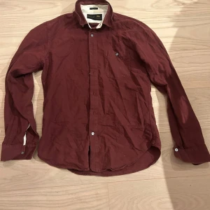 Vinröd skjorta från Woolrich - Snygg vinröd skjorta från Woolrich med klassisk krage, bröstficka och knäppning framtill. Skjortan är långärmad och tillverkad i mjuk bomull, perfekt för dig som gillar stilrena plagg med enkel design.