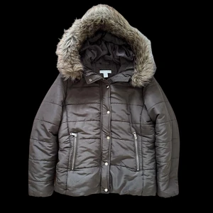 Grå pufferjacka med fuskpäls - Snygg grå pufferjacka med huva och fuskpäls runt kanten. Jackan har quiltad design, dragkedja och knappar framtill samt två fickor med dragkedja. Perfekt för kalla dagar och ger en cool vibe till din vinterstil.