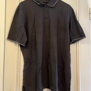 Mörkgrå pikétröja från Structure XL - Snygg mörkgrå pikétröja från Structure i storlek XL. Tröjan är i 100% pima bomull och har klassisk krage med knappar och vita detaljer längs krage och ärmslut. Perfekt för en clean och stilren look.