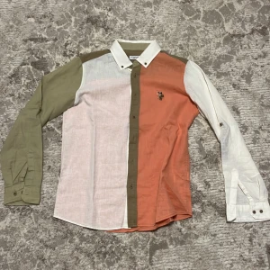 Flerfärgad skjorta från U.S. Polo Assn. - Snygg skjorta från U.S. Polo Assn. med blockfärger i olivgrönt, vitt och orange. Klassisk krage, broderad logga på bröstet och knappar framtill. Tillverkad i linne för en skön och luftig känsla. Perfekt för dig som gillar att sticka ut med färg och stil.