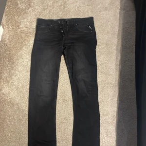 Svarta Replay jeans Anbass - Svarta jeans från Replay, modell grover. Klassisk femficksdesign med knappgylf och diskret Replay-logga vid fickan. Jeansen har en slim passform och raka ben, tillverkade i stretchigt bomullsmaterial för extra komfort.storlek 32