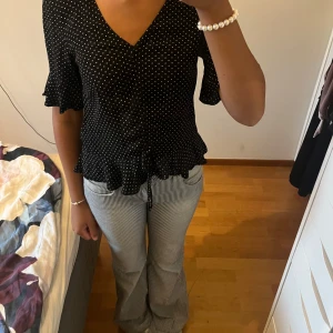 Svart prickig blus från H&M - Svart blus från H&M med vita prickar, v-ringning och volangkant nertill. Korta ärmar med volang och snörning framtill. 