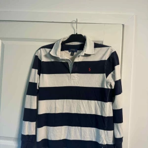 Randig pikétröja från Polo Ralph Lauren - Säljer en klassisk långärmad pikétröja från Polo Ralph Lauren med breda marinblå och vita ränder. Tröjan har krage, knappar vid halsen och den ikoniska röda loggan broderad på bröstet. Perfekt för en sportig och stilren look. XS-S