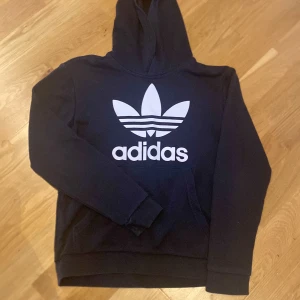 Svart Adidas hoodie med stor logga - Svart hoodie från Adidas med stor vit Trefoil-logga och text på bröstet. Klassisk huva och magficka, långärmad modell i mjukt material. Perfekt för en sportig och avslappnad stil.