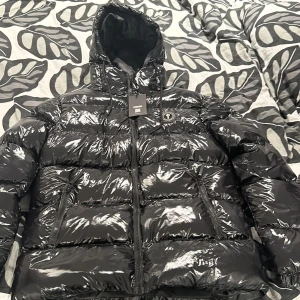 Svart pufferjacka från Trapstar - Snygg svart pufferjacka från Trapstar med glansig finish och huva. Jackan har dragkedja och märkets logga på bröstet. Perfekt för kyliga dagar.