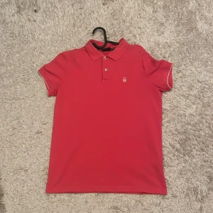 Röd pikétröja från Sail Racing S - Riktigt snygg Rosa pikétröja från Sail Racing med klassisk krage och två knappar framtill. Tröjan har vit logga på bröstet och diskreta vita detaljer vid ärmsluten. Perfekt för en fräsch och sportig look.