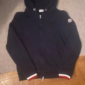 Mörkblå hoodie från Moncler - Säljer en mörkblå hoodie från Moncler med dragkedja och huva. Tröjan har Moncler-logga på ärmen och ribbade muddar med röd och vit detalj. Perfekt för dig som gillar stilrena och exklusiva plagg.