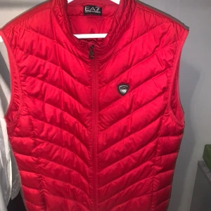  EA7 väst - Snygg röd dunväst från Emporio Armani EA7 med quiltad design och dragkedja framtill. Västen har en stilren look med EA7-logga på bröstet och hög krage. Perfekt för lager-på-lager och streetstyle.