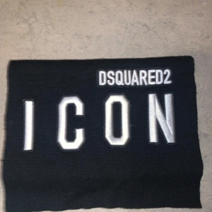halsduk från Dsquared2 ICON - Svart halsduk från Dsquared2 med stor, vit brodyrtext 'ICON' och Dsquared2-logga. Halsduken är stickad och har en clean, rektangulär form som ger en modern vibe. Perfekt accessoar för att lyfta din outfit med en exklusiv touch.
