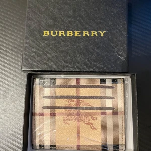 Burberry rutig plånbok i skinn - Snygg plånbok från Burberry i klassiskt rutigt mönster med beige, bruna och svarta toner. Tillverkad i skinn och har flera kortfack på framsidan. Plånboken har Burberrys ikoniska logga med riddare tryckt på framsidan. Perfekt accessoar för dig som gillar stilren design.