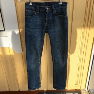 Blå jeans från Nudie Jeans, 27/30 - Säljer nu ett par snygga blå jeans från Nudie Jeans i klassisk femficksmodell. Jeansen har orangea kontrastsömmar, mellanblå tvätt och raka ben. Tillverkade i slitstarkt bomullstyg med Nudie-logga på bakfickan och patch i midjan. Perfekta för en avslappnad och stilren look.