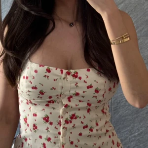 Blommig toop med knappar - Supersöt topp med blommigt mönster i rött och grönt på en ljus, nästan vit bakgrund. Klänningen har knappar framtill och en figurnära passform som framhäver midjan. Perfekt för dig som gillar romantisk och feminin stil.