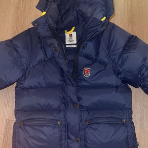 Blå dunjacka från Fjällräven - Säljer en blå dunjacka från Fjällräven med huva och klassisk logga på bröstet. Jackan har dragkedja, tryckknappar och stora fickor framtill. Perfekt för kalla dagar och har snygga gula detaljer på insidan av kragen.