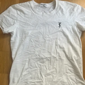 Saint Laurent t shirt  - Vit t-shirt från Saint Laurent med klassisk rund hals och svart YSL-broderi på bröstet. Enkel och stilren design i mjuk bomull, perfekt till jeans eller shorts. Kortärmad modell med normal passform. Storlek M men liten i storleken passar S, Inte använd så mycket 