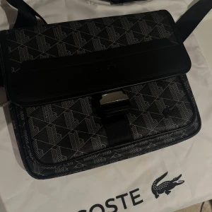 Svart axelväska från Lacoste med mönster - Snygg svart axelväska från Lacoste med geometriskt mönster och diskret logga framtill. Väskan har justerbar axelrem, lock med spänne och detaljer i skinn och syntet. Perfekt för att bära dina viktigaste grejer med stil.