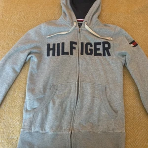 Grå hoodie från Tommy Hilfiger XS - 🚨Pris kan diskuteras 🚨Snygg grå hoodie från Tommy Hilfiger i storlek XS. Tröjan har dragkedja, huva med vita snören och stora mörkblå bokstäver på bröstet. Klassisk Hilfiger-flagga på ärmen och två fickor framtill. Perfekt för en chill och sportig look.