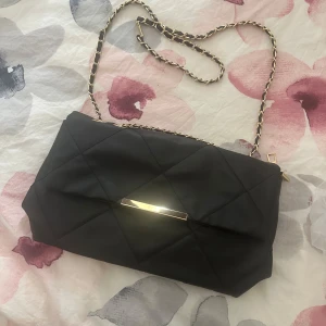 Svart quiltad axelväska med guldkedja - Stilren svart axelväska med quiltat mönster och guldfärgade detaljer. Väskan har en kedjerem med inslag av svart material och ett elegant guldfärgat spänne framtill. Perfekt storlek för det viktigaste och med flera fack inuti för bra organisering.