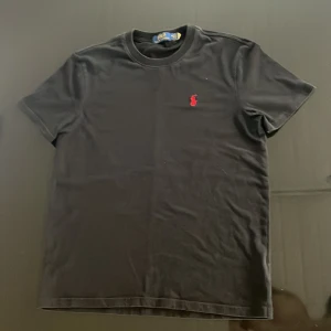 Svart t-shirt från Polo Ralph Lauren - Svart t-shirt från Polo Ralph Lauren med klassisk rund hals och korta ärmar. Står M men sitter som en S. På bröstet finns den ikoniska röda Polo-loggan broderad. T-shirten är gjord i mjuk bomull och har en clean, stilren look som funkar till allt.