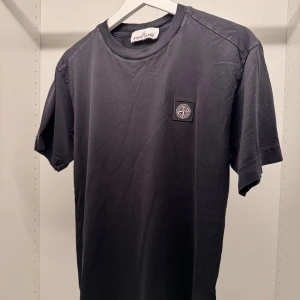 Svart t-shirt från Stone Island - Svart t-shirt från Stone Island med klassisk rund hals och korta ärmar. T-shirten har en stilren look med Stone Island-logga på bröstet. Tillverkad i mjuk bomull för skön passform och enkel stil.
