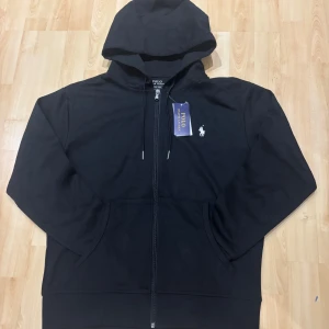 Svart hoodie från Polo Ralph Lauren - Svart hoodie från Polo Ralph Lauren med dragkedja framtill och klassisk huva med snörning. Tröjan har en vit broderad logga på bröstet. Prfekt för en chill och stilren look.