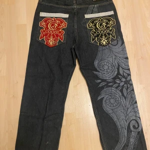 COOGI jeans med brodyr och mönster - Unika svarta jeans från COOGI med stora broderade loggor i rött och gult på bakfickorna och detaljerad grå mönsterprint längs ena benet. Guldigt broderi på myntfickan och klassisk femficksdesign. Raka ben och cool streetwear-vibe.