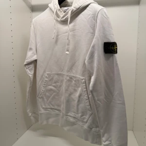 Vit hoodie från Stone Island - Vit hoodie från Stone Island med klassisk patch på vänster ärm. Tröjan har huva med snörning, stor magficka och långa ärmar. Tillverkad i mjuk bomull, perfekt för en clean och stilren look.