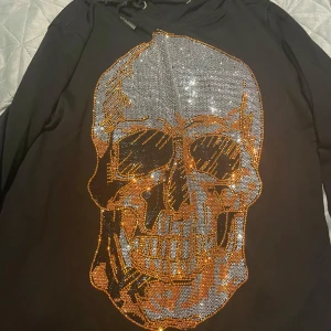 Svart hoodie med döskalle från Philipp Plein - Svart hoodie från Philipp Plein med stor glittrig döskalle framtill i silver och guld. Tröjan har huva med snörning och är tillverkad i mjuk bomull. Snygg detalj med logotyp på huvan och insidan. Perfekt för dig som gillar statement-plagg.