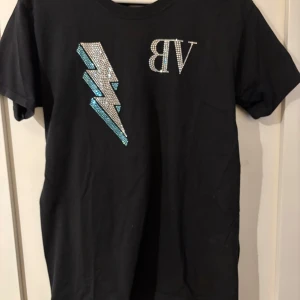 Svart t-shirt med blixt - Svart t-shirt i bomull med korta ärmar. På framsidan finns en stor blixt i silverfärgade och blå strasstenar samt bokstäverna 'BV' i strass. Snygg och fet tröja.