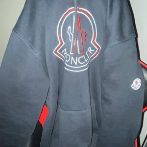 Svart Moncler hoodie med stor logga - Svart hoodie från Moncler i storlek XXL, men passar också mig och jag har S. Det är sitter också en stor broderad logga i vitt och rött på bröstet samt Moncler-märke på ärmen. Klassisk känguruficka framtill och snörning i huvan. Perfekt för dig som gillar streetwear och exklusiva märken.