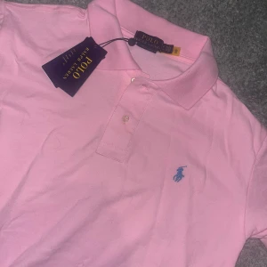 Rosa pikétröja från Polo Ralph Lauren - Snygg rosa pikétröja från Polo Ralph Lauren med klassisk krage, knappar framtill och blå broderad logga på bröstet. Tillverkad i mjuk bomull och har korta ärmar. Perfekt för en clean och stilren look.