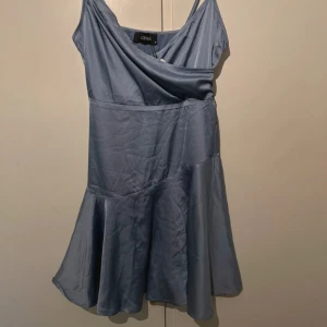 Ljusblå omlottklänning från Cider - Supersöt ljusblå kortklänning från Cider med omlott-detalj upptill och tunna axelband. Klänningen har en lätt glansig finish och är sydd i ett mjukt, följsamt material som ger en snygg siluett. Perfekt för dig som vill ha en stilren men ändå unik look.