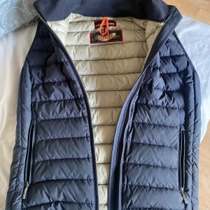 Parajumpers dunväst navy S - Snygg dunväst från Parajumpers i mörkblått med ljus insida. Västen har quiltad design, två dragkedjefickor och en orange detalj vid kragen. Perfekt för lager-på-lager och riktigt lätt att bära.