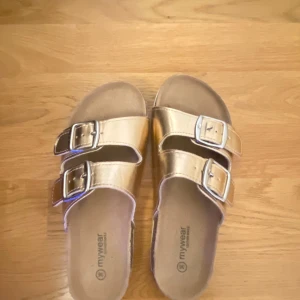 Guldiga sandaler från Mywear, stl 36 - Snygga sandaler i metallic guld från Mywear med två breda remmar och stora silverspännen. Fotbädden är i skinn och sulan är beige med mönster undertill. 