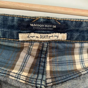 Maison Scotch blå jeans med slitningar - Snygga blå jeans från Maison Scotch med slitna detaljer och knappar i gylfen. Jeansen har klassisk femficksmodell och rutigt innerfoder i midjan. Perfekt för dig som gillar en avslappnad och trendig look.