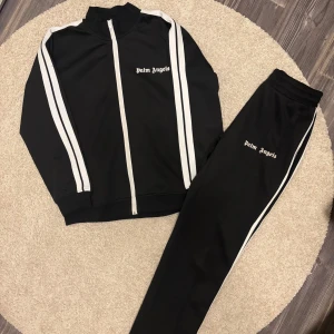 Palm Angels svart track jacket - Svart track jacket från Palm Angels med vita ränder längs ärmarna och vit dragkedja. Klassisk sportig look med hög krage och logga på bröstet. Materialmix av bomull och polyester, perfekt för en streetwear vibe.