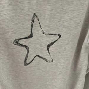 Grå hoodie med stjärna och text - Gråmelerad hoodie med huva och snörning. På bröstet står det 'AMOUR' i svart, sliten stil och på ryggen finns en stor svart stjärna. Mjuk och skön, perfekt för chill dagar. Långärmad och med klassisk passform.