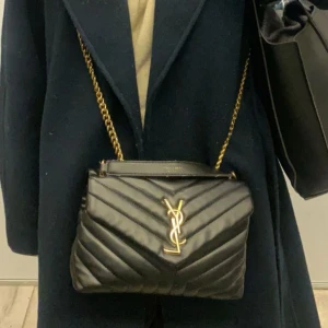 Svart quiltad YSL axelväska i skinn - Säljer en svart quiltad axelväska i skinn med guldiga kedjor och YSL-logga framtill. Väskan har elegant V-mönster och en lyxig känsla, perfekt för dig som vill sticka ut med en klassisk designerbag. ❤️Den är köpt på plick för 2000 så vet inte ifall den är äkta men den ser äkta ut. !!Kan gå ner i pris, kom även med prisförslag om ni inte tycker att priset som är satt är bra!!❤️