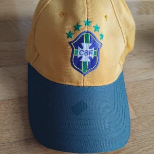 Gul Brasil keps med CBF-logga - Snygg gul keps med grön skärm och broderad CBF-logga framtill, samt texten 'Brasil' broderad baktill. Kepsen har justerbar rem och är tillverkad i bomull. Perfekt för dig som älskar fotboll och vill visa support för Brasilien.