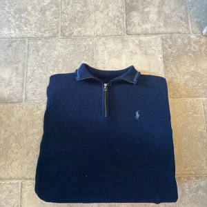 Marinblå stickad tröja Polo Ralph Lauren - Säljer en marinblå stickad tröja från Polo Ralph Lauren med halv dragkedja och klassisk polospelare broderad på bröstet. Tröjan har ribbade muddar, lång ärm och en snygg krage med diskreta ränder. Perfekt för dig som gillar stilren och tidlös design.